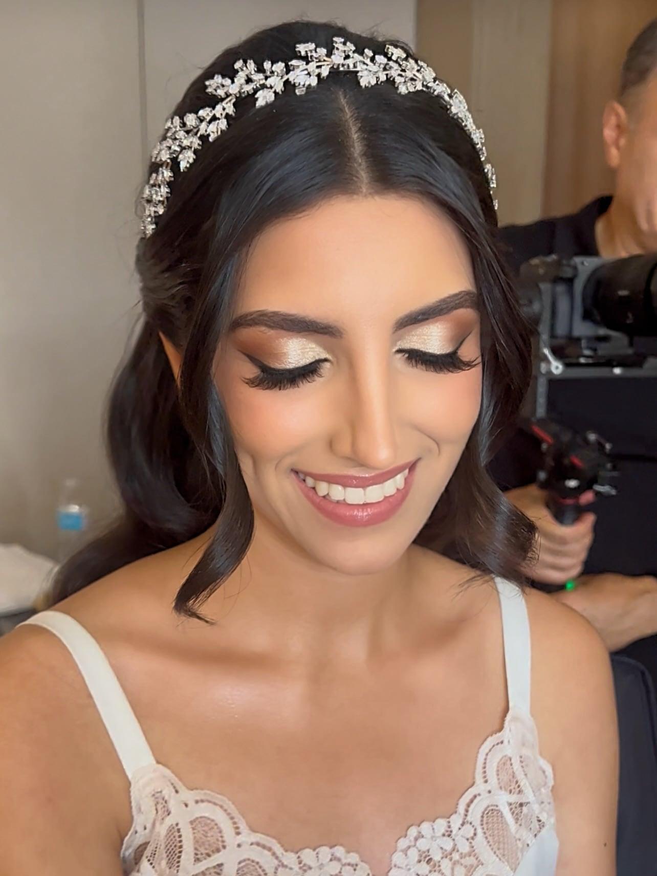 Smiling bride with bronze shimmer eyes, full lashes, and a crystal headpiece while a videographer films