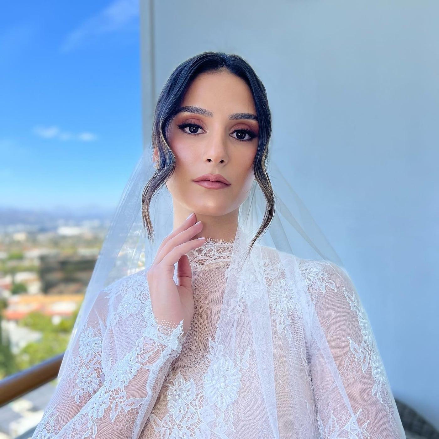 Bride in a lace gown and sheer veil with defined eyes and a soft rose lip, city view behind her