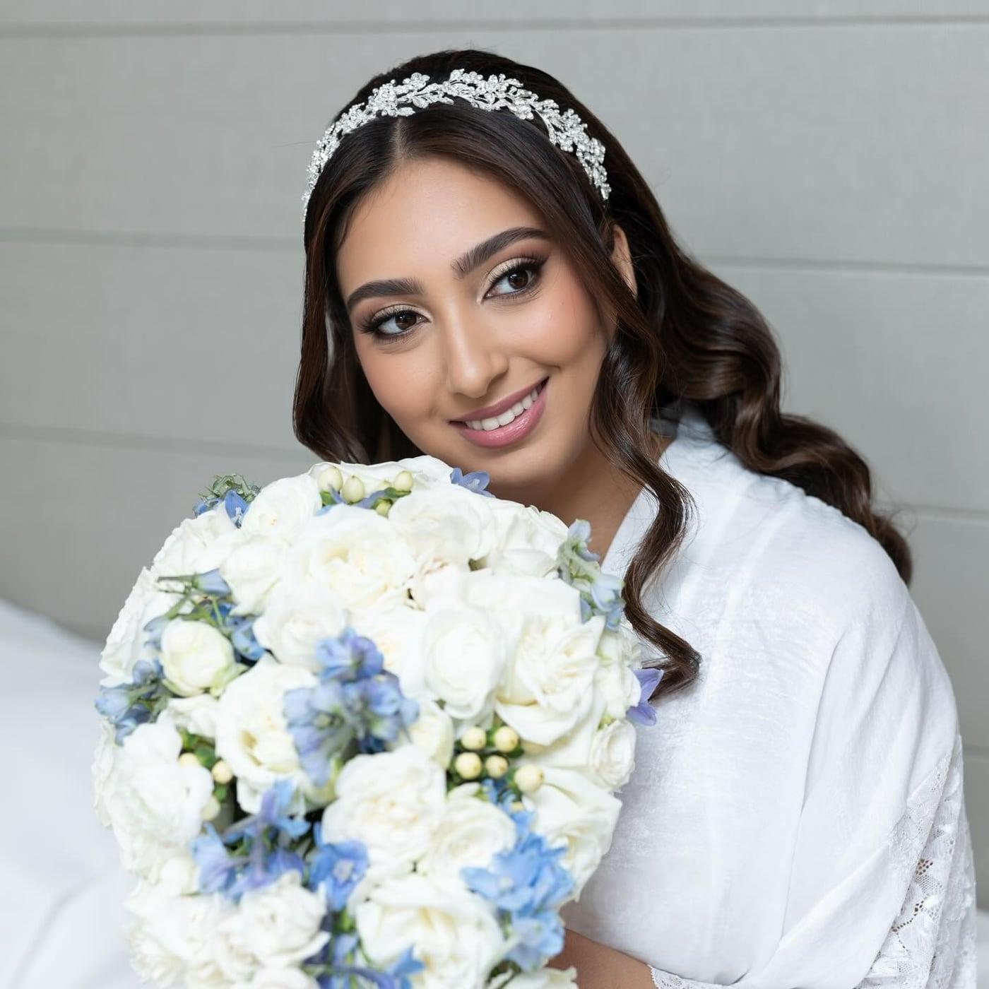 Smiling bride with glam makeup and a crystal headpiece holding a white and blue bouquet