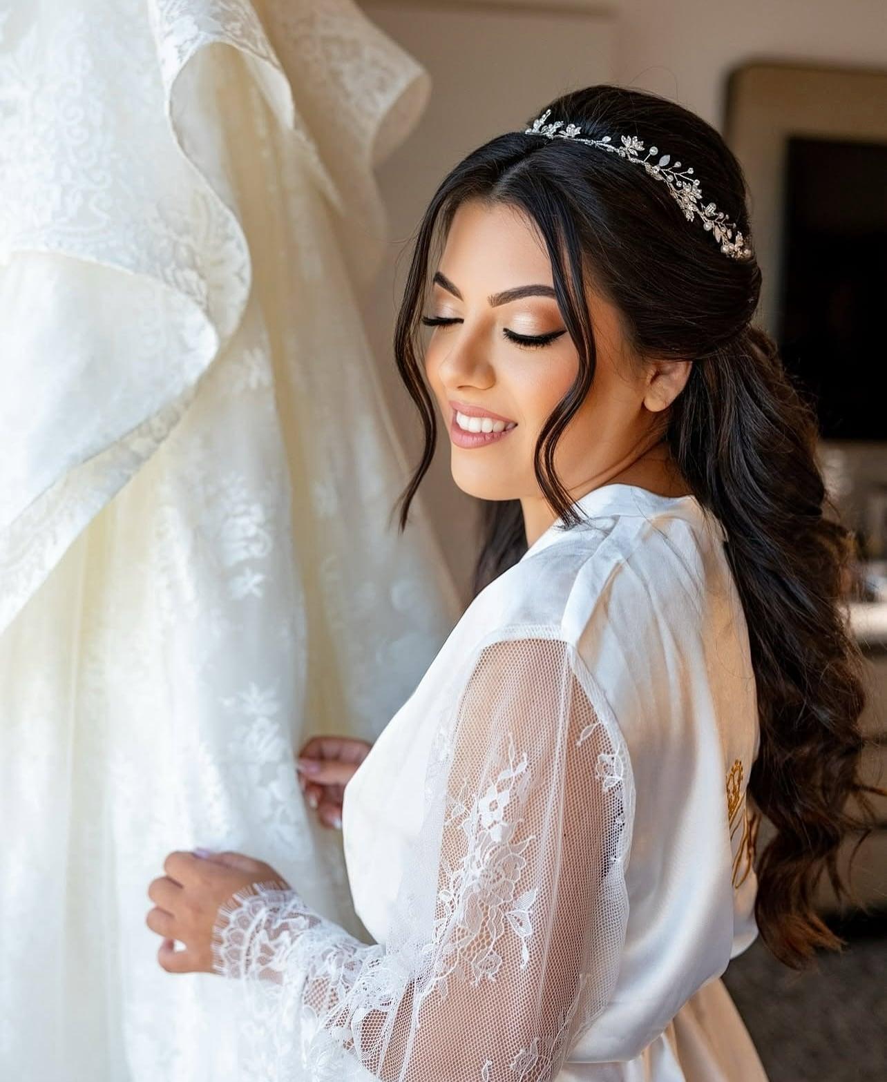Bride in a lace robe with soft glam makeup and a crystal hair vine, touching her wedding gown