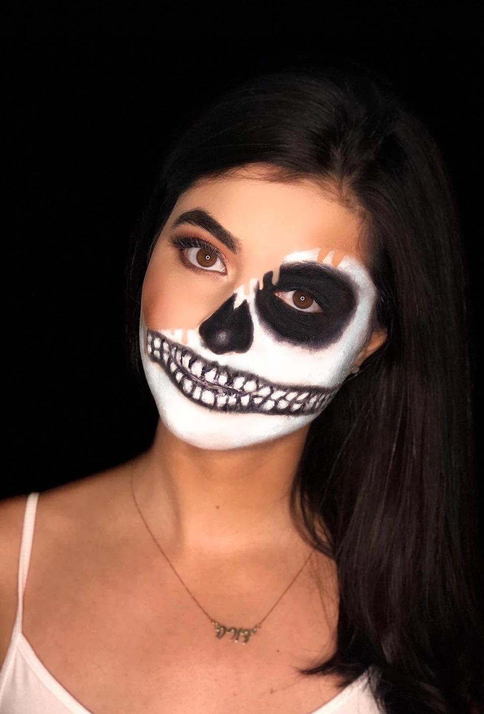 Half-face skull makeup: one side painted with a white base, a blacked-out eye socket, a dark nose cavity and teeth drawn across the lips, the other side left bare